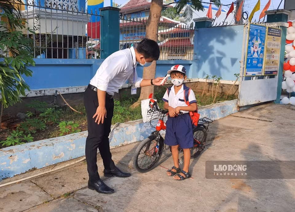 A boy cycles to the school by himself. He has his body temperature measured and is guided to use a bottle of hand sanitizer before entering the school.